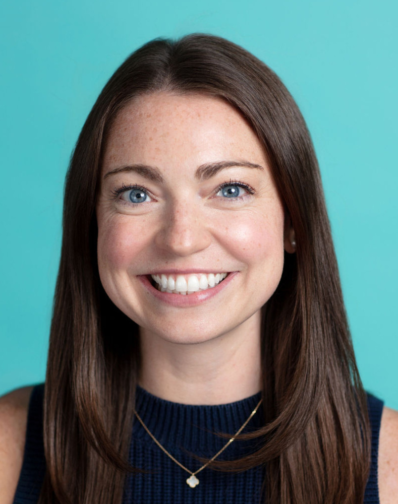 A woman with long brown hair wearing a navy sleeveless top and a necklace, smiling in front of a teal background.