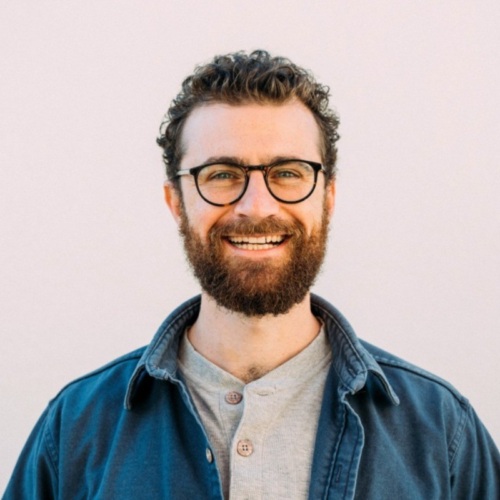 A bearded man with glasses is smiling at the camera. He is wearing a light gray shirt and a blue jacket against a plain, light-colored background.