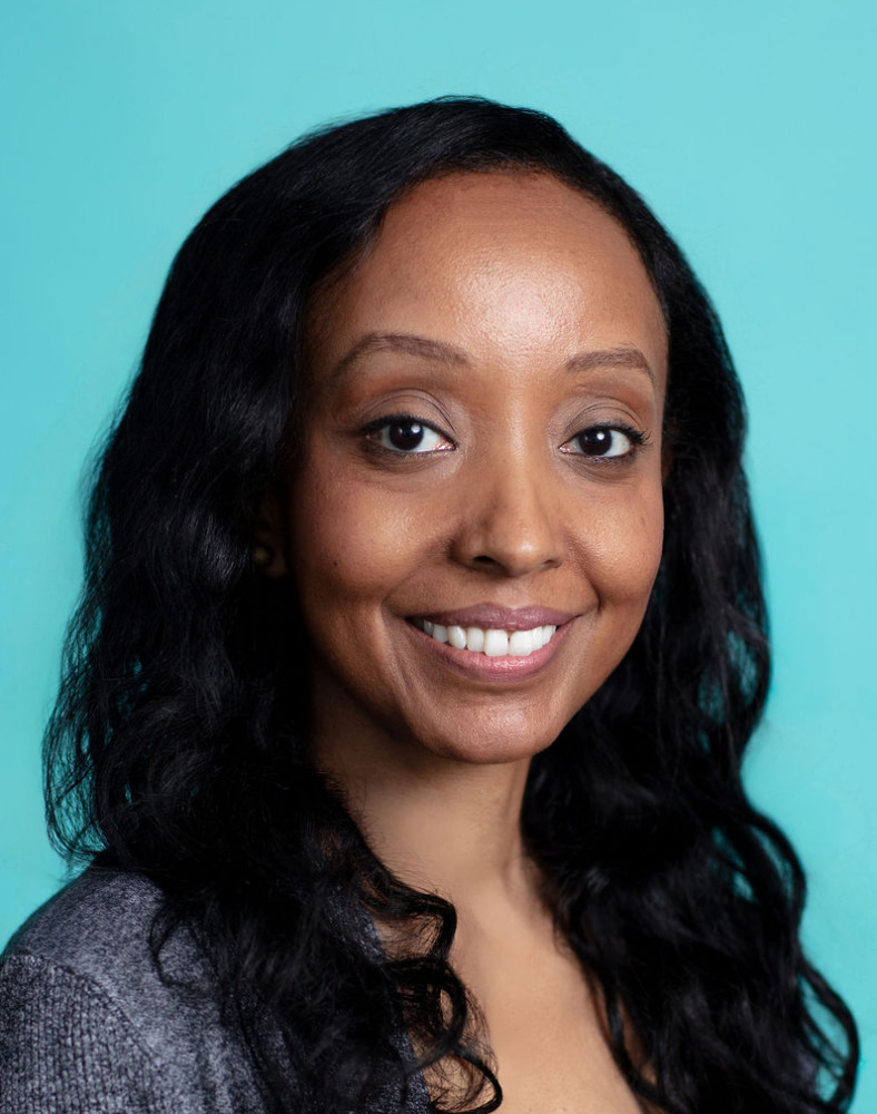Woman with long black hair wearing a gray cardigan and black top, smiling, posed against a plain turquoise background.