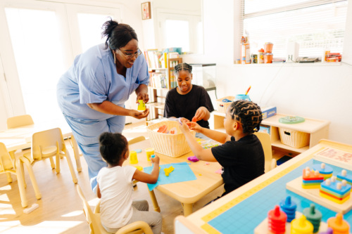 A caregiver in scrubs interacts with three children at a table, helping them play with toys and modeling clay in a brightly lit classroom.