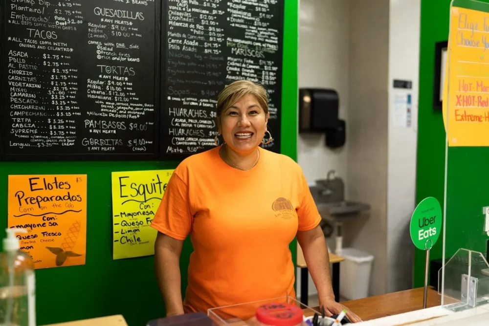 A woman wearing an orange shirt stands smiling behind a counter in a restaurant, with menu boards and signs displayed on the wall behind her.