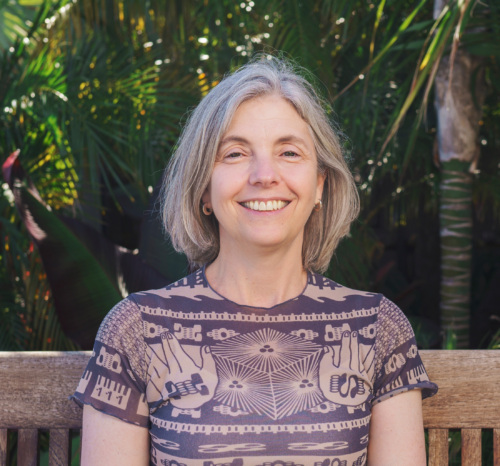 A woman with gray hair sits on a wooden bench outdoors, smiling, with green foliage in the background.