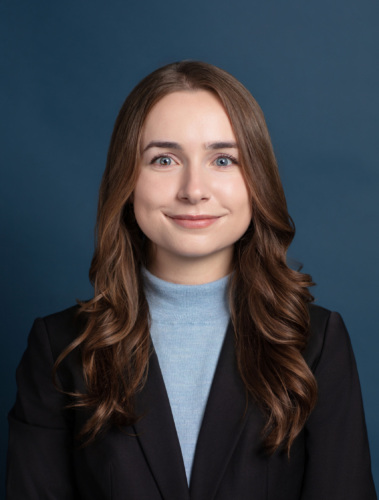 A young woman with long brown hair, wearing a black blazer and light blue turtleneck, poses in front of a plain dark blue background.