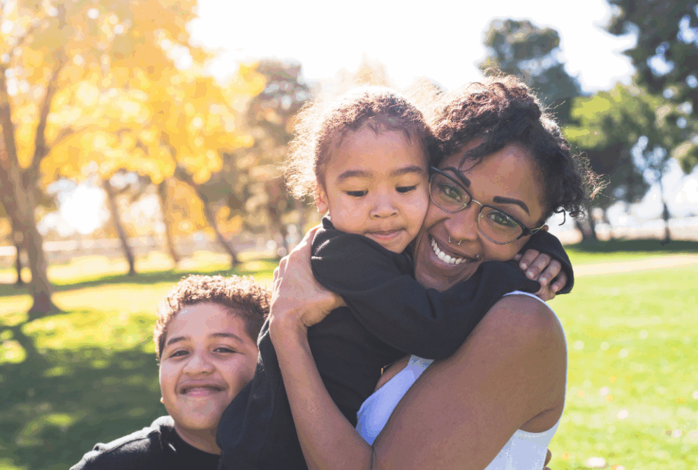 A woman wearing glasses hugs a young girl while a smiling boy stands beside them in a sunny park with green grass and trees.