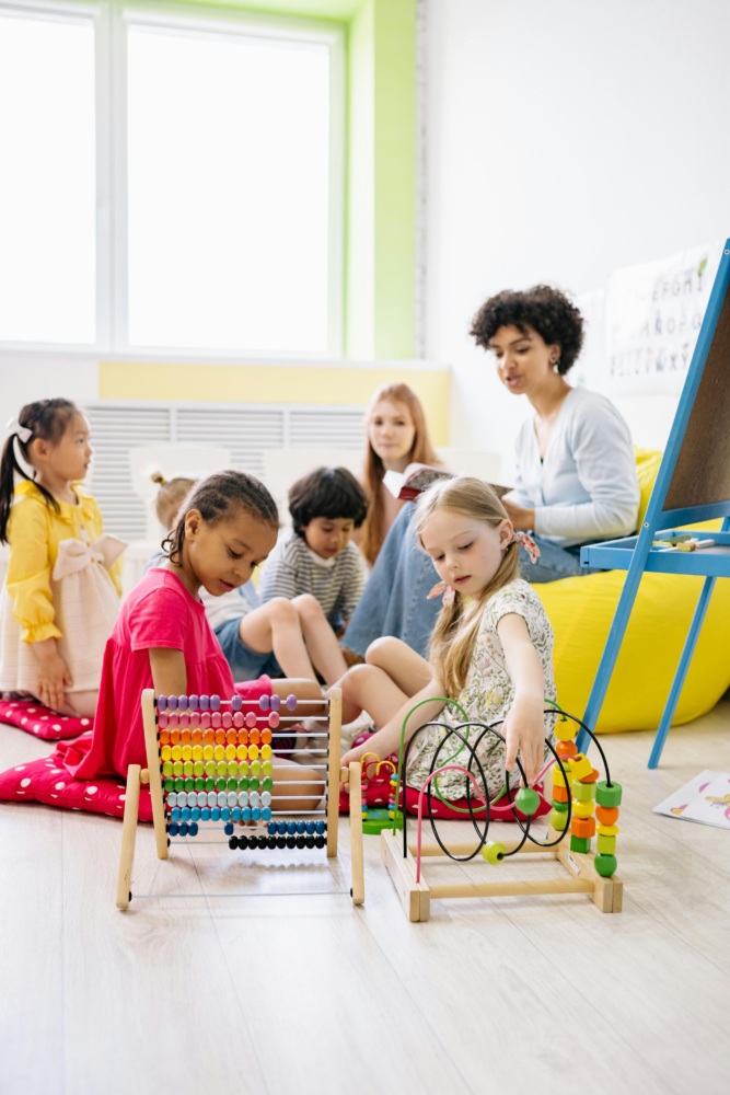 Several young children play with educational toys on the floor while an adult sits nearby in a brightly lit classroom.
