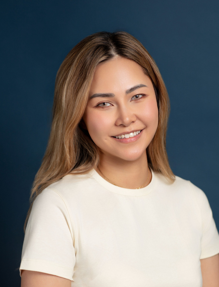 A woman with long light brown hair wearing a cream-colored shirt smiles at the camera against a dark blue background.
