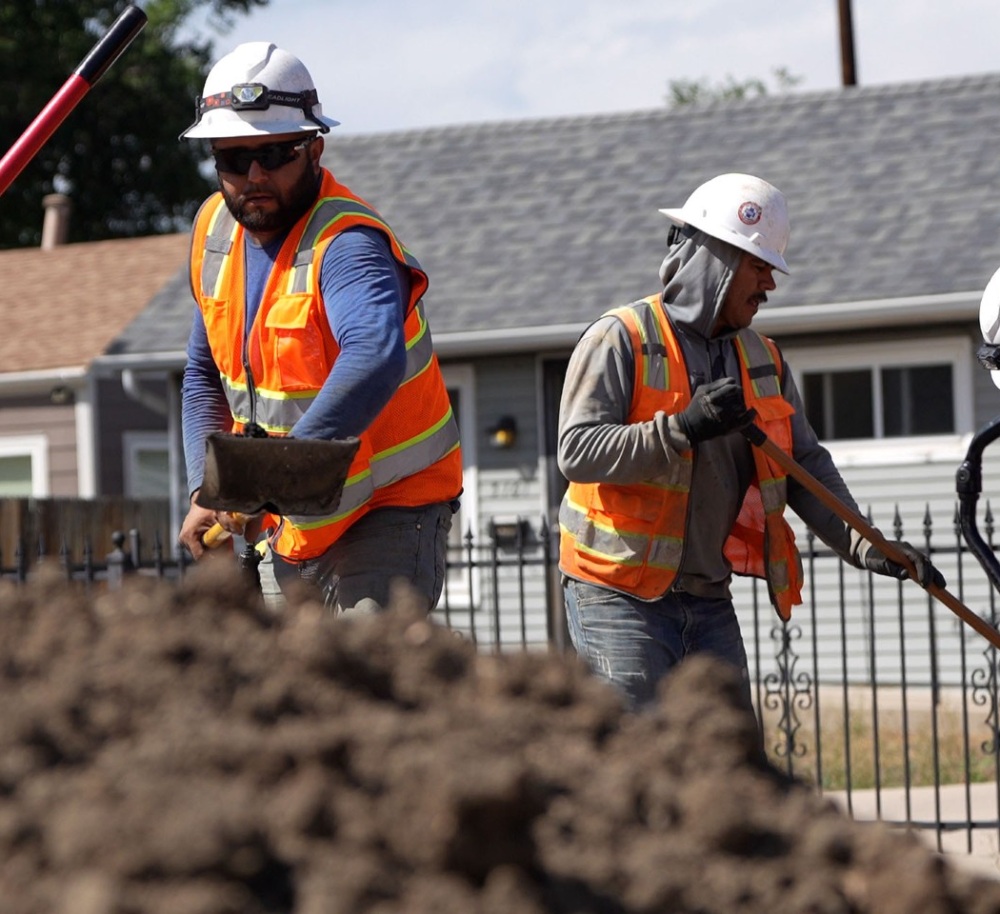 Four construction workers in safety gear and reflective vests work with shovels near a pile of dirt in a residential area.