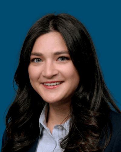 A woman with long dark hair, wearing a dark blazer and light shirt, smiling in front of a solid blue background.