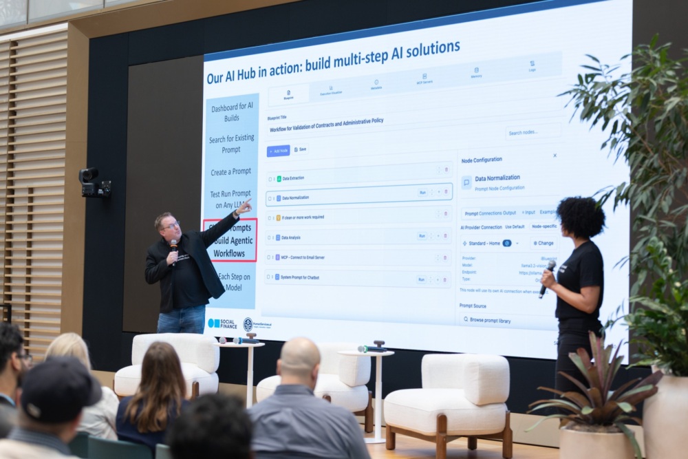 A man presents AI workflow steps on a large screen to an audience, while a woman stands by with a microphone in a modern conference room.