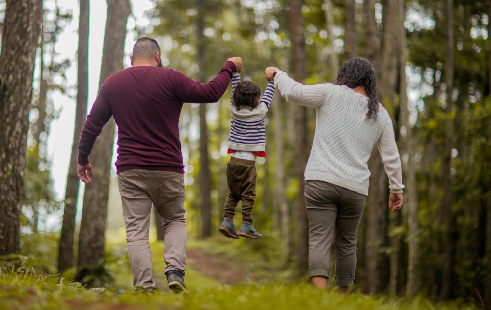 Two adults walk hand-in-hand with a child through a forest, holding the child's hands and lifting them off the ground as they walk along a dirt path.