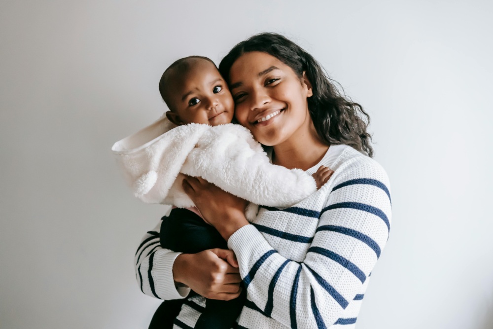 A woman in a striped sweater smiles while holding a baby in a white fuzzy jacket against a plain, light-colored background.