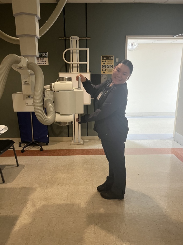A person in medical scrubs operates an X-ray machine in a clinical setting with caution signs visible on the wall.