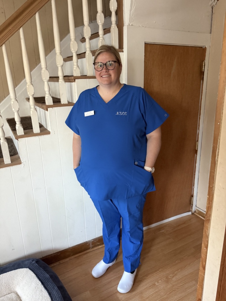 A person wearing blue medical scrubs and white shoes stands indoors near a staircase and wooden door, with hands in pockets.