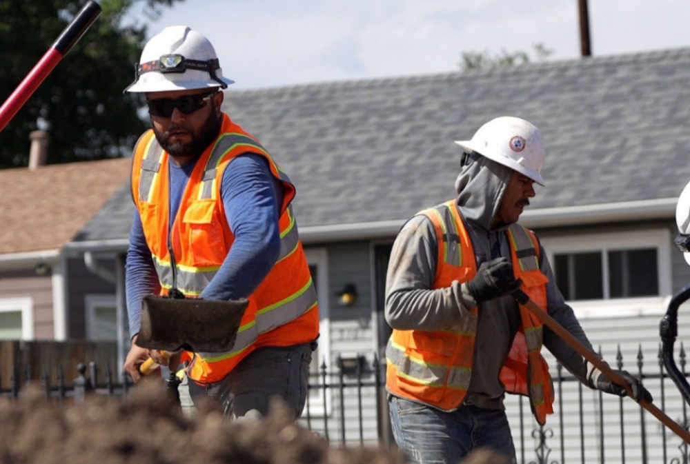 Two construction workers in safety gear and orange vests shovel dirt at a construction site in front of residential houses.