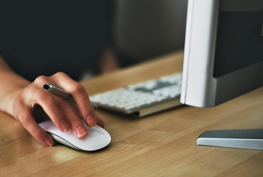 A person uses a computer mouse next to a keyboard and monitor on a wooden desk.