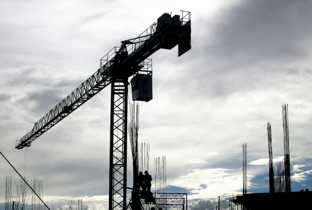 A construction crane towers over a building site with two workers standing on scaffolding against a cloudy sky.
