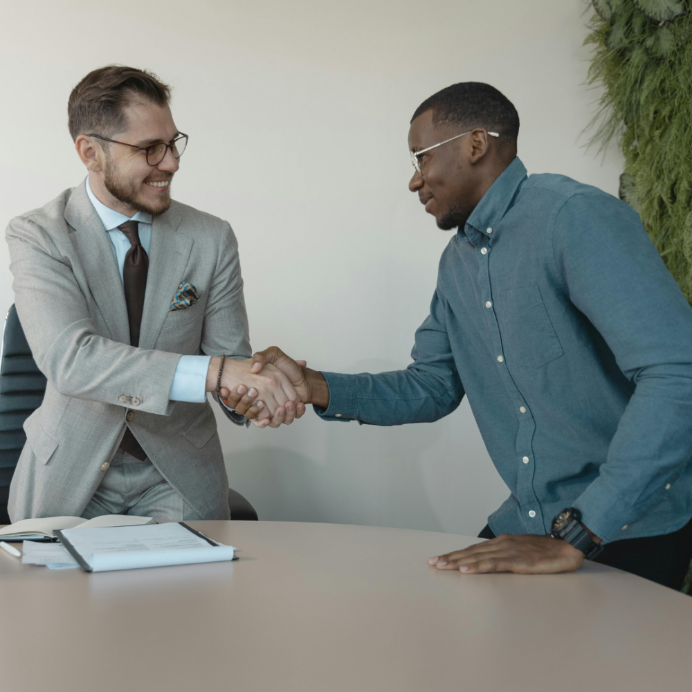 Two men shake hands across a desk with documents and a pen, one wearing a suit and the other in a casual shirt, in a modern office setting.
