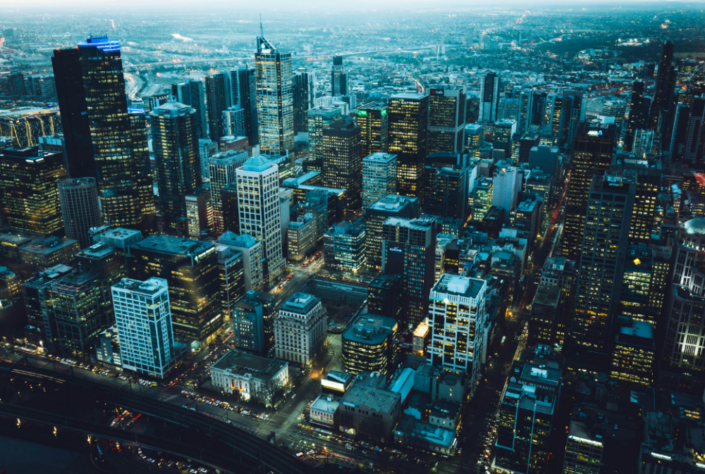 Aerial view of a city at dusk showing numerous high-rise buildings with lights on, busy roads, and a sprawling urban landscape extending into the distance.