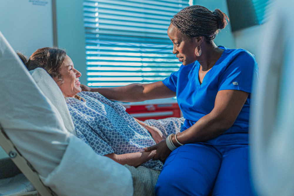 A nurse in blue scrubs sits beside a patient in a hospital bed, holding her hand and offering support in a brightly lit hospital room.