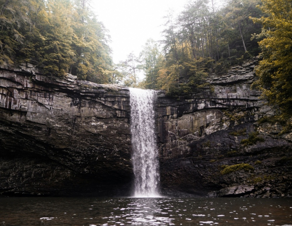 A tall, narrow waterfall flows over a rocky cliff into a calm pool, surrounded by forested trees under an overcast sky.