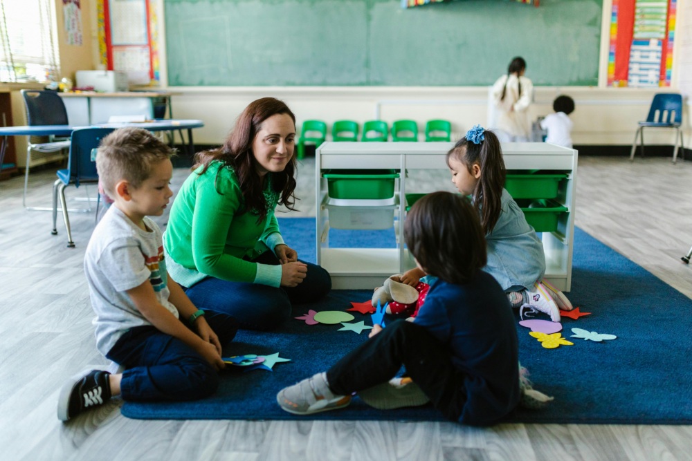 A teacher and three young children sit on a blue rug in a classroom, interacting with colorful paper shapes. A chalkboard and desks are visible in the background.