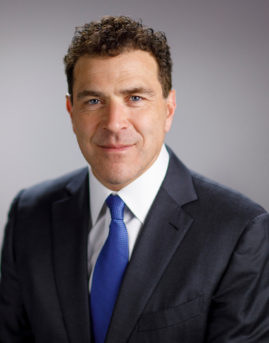 A man in a dark suit, white shirt, and blue tie poses against a plain gray background, looking directly at the camera.
