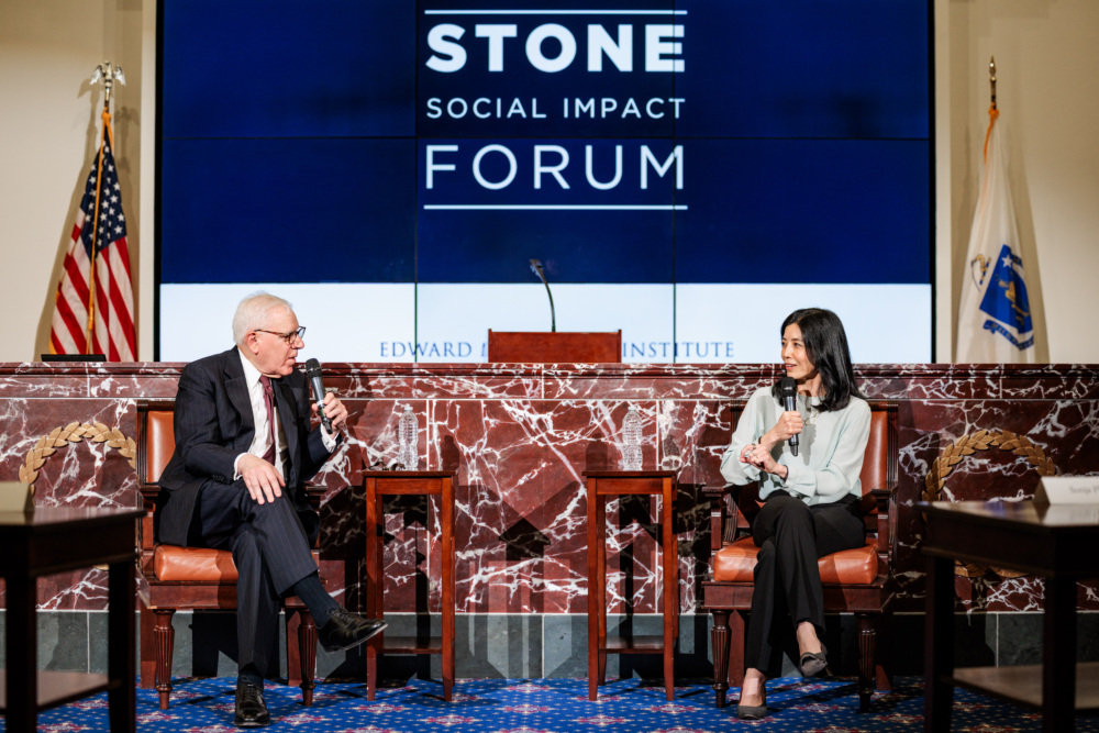 Two people sit on stage having a discussion with microphones at the Stone Social Impact Forum, with a large screen and flags in the background.