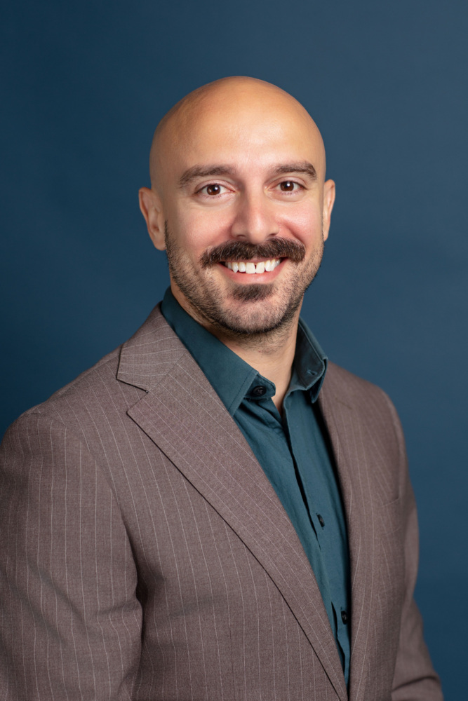 A smiling man with a bald head and mustache, wearing a brown pinstripe suit jacket and teal shirt, poses in front of a plain dark blue background.