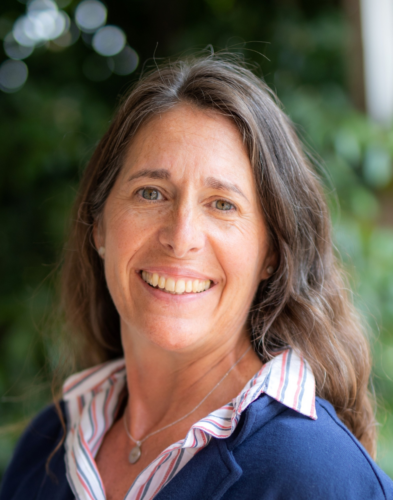 A woman with long brown hair, wearing a blue blazer over a striped shirt, smiles at the camera with greenery in the background.