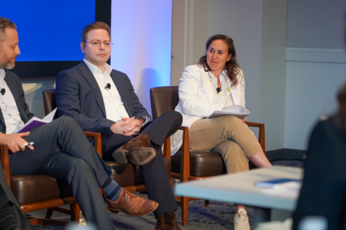 Two men in business attire sit and participate in a panel discussion; one gestures while speaking, and the other listens attentively.