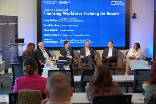 Five people in business attire sit on a stage panel, engaged in discussion, with microphones clipped to their clothing and notes in hand. A podium and screen are visible in the background.