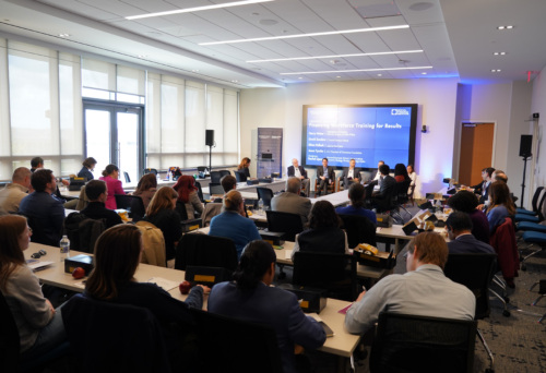 People attend a panel discussion in a modern conference room. Five panelists sit at the front, and a presentation screen displays the event topic, "Promoting Workforce Training for Results.