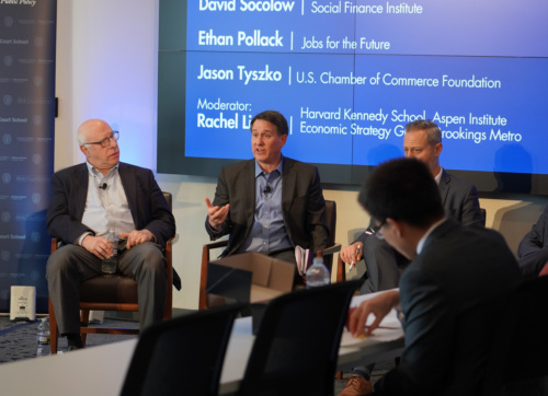 Four men sit on a panel in discussion, with a moderator and speakers' names displayed on a large screen behind them. One panelist gestures as he speaks.