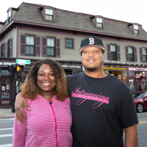 Two people stand close together and smile on a city street in front of a brown building with shops and apartments.
