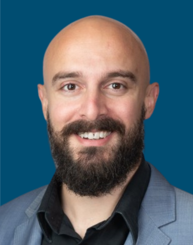A man with a bald head and full beard wearing a gray suit jacket and black shirt, smiling in front of a solid blue background.