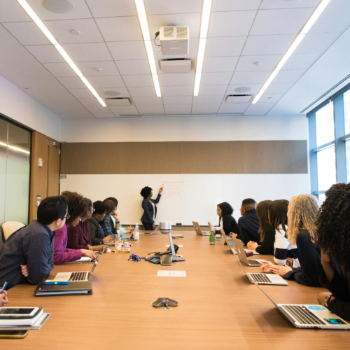 A person stands at a whiteboard giving a presentation to a group of people seated around a conference table with laptops.