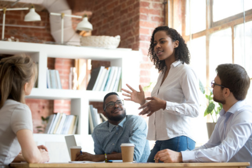 Four people in a modern office sit around a table having a discussion. One woman stands and speaks while the others listen attentively.