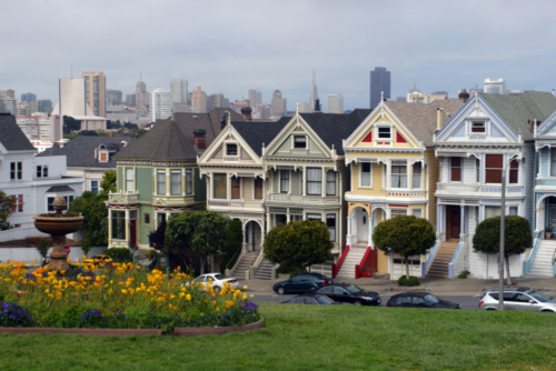 Victorian-style houses known as the Painted Ladies stand in a row with a city skyline in the background and a grassy park with flowers in the foreground.