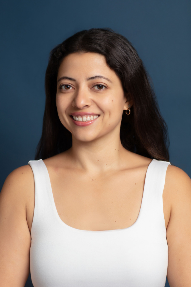 Woman with long dark hair wearing a white sleeveless top, smiling softly, posed in front of a plain dark blue background.