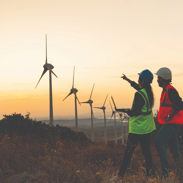 Two people in safety gear stand in a field at sunset, observing and discussing several wind turbines in the background.