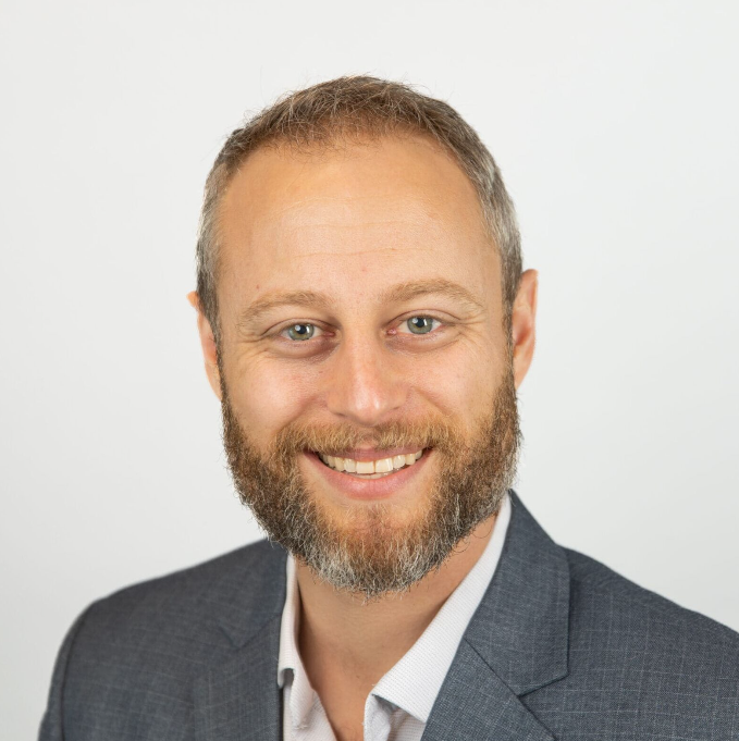 A man with short hair and a beard, wearing a gray suit jacket and light-colored shirt, smiles at the camera against a plain white background.