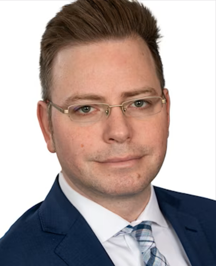 A man with short brown hair and glasses wearing a blue suit, white shirt, and patterned tie, facing the camera against a white background.