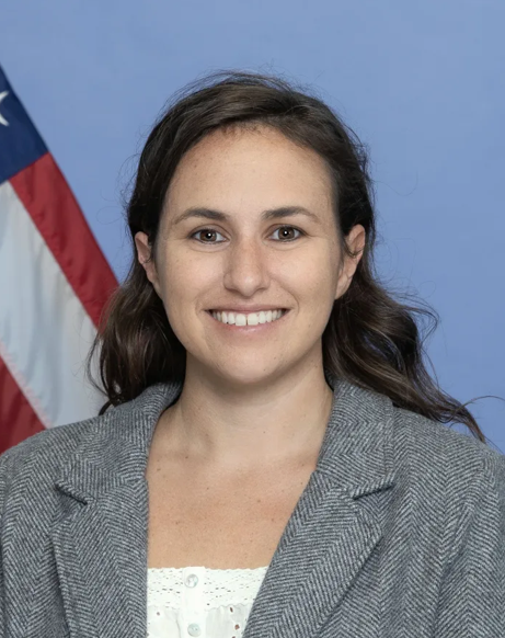 A woman with long brown hair, wearing a grey blazer and white top, smiles in front of a blue background and a partially visible American flag.