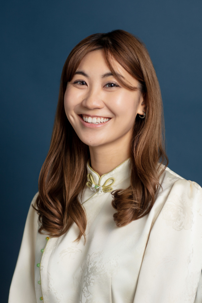 A woman with long brown hair wearing a cream-colored traditional blouse smiles in front of a plain dark blue background.