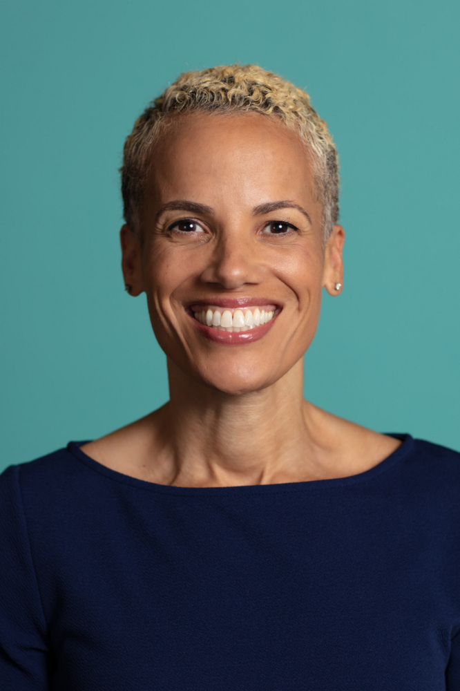 A woman with short, curly blonde hair smiles at the camera, wearing a dark blue top against a teal background.