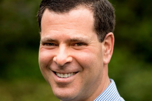 A man with short dark hair smiles at the camera outdoors, wearing a blue checkered shirt; green blurred background.