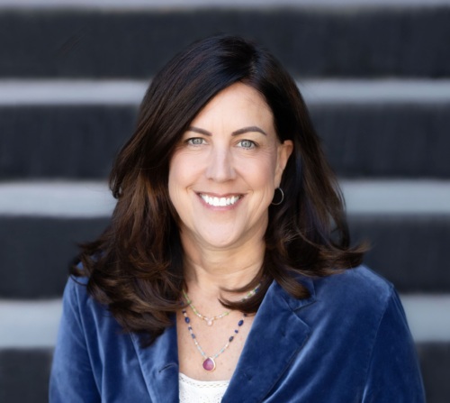 Woman with long dark hair in a blue blazer smiling, sitting outdoors in front of gray stone steps.