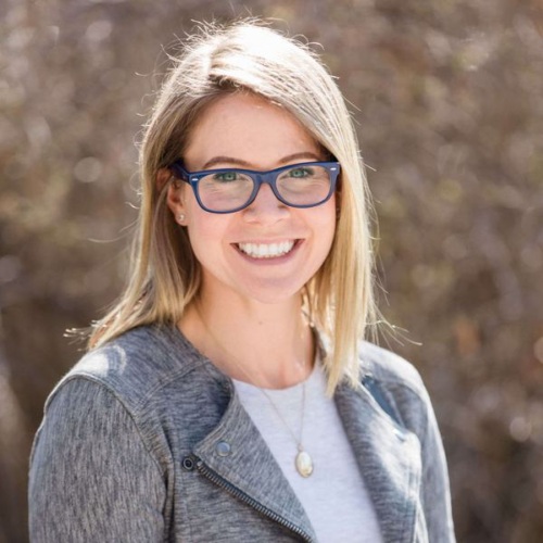 A woman with straight blonde hair and blue glasses smiles at the camera, wearing a grey jacket and a white top, with a blurred outdoor background.