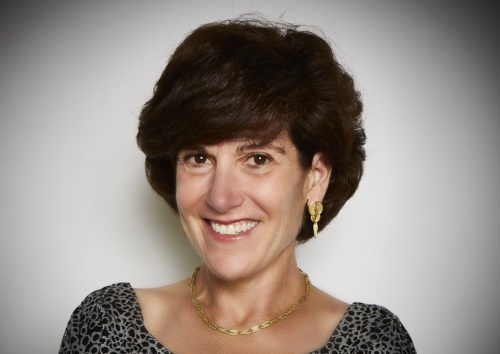 A woman with short dark hair wearing a patterned top, gold necklace, and gold earrings, smiling at the camera against a plain background.