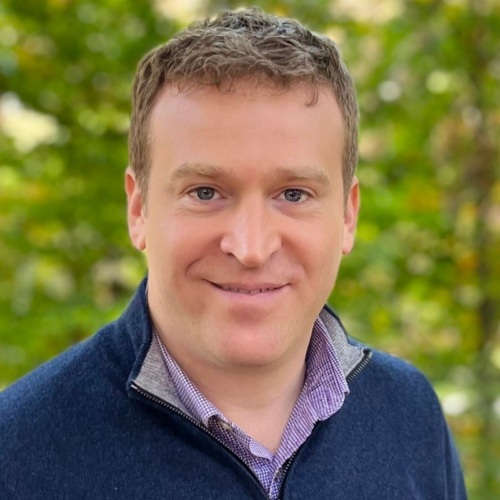 A man with short light brown hair, wearing a navy sweater over a collared shirt, smiles at the camera with a blurred green background.